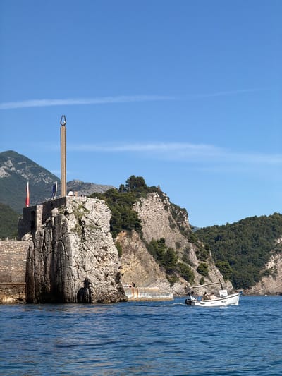 In Petrovac centar, Montenegro, a bright, clear blue sky is visible above a rugged coastal landscape. In the foreground, the deep blue sea ripples gently. A small white motorboat with a canopy is navigating the water, carrying several passengers. The boat has a visible registration or nameplate. To the left of the boat, a rocky cliff face rises steeply, topped by a stone structure with two flags – one red and one blue. Dominating the top of this structure is a tall, slender obelisk-like monument. Beyond the cliff, lush green trees cover the slopes of distant mountains, also under the clear blue sky. The overall scene depicts a serene coastal environment under sunny conditions.