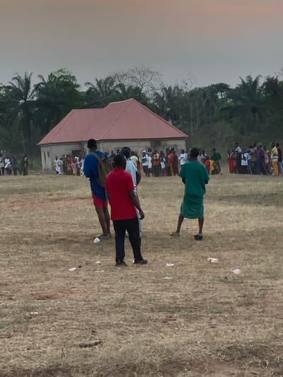 Several individuals stand on dry grass facing a building with a red roof. One person is dressed in a green uniform, and a group is wearing red and blue shirts with dark pants. A crowd of people stands in the background behind the building and near some trees. Litter is scattered on the dry grass. The sky is partly cloudy with a muted color palette.