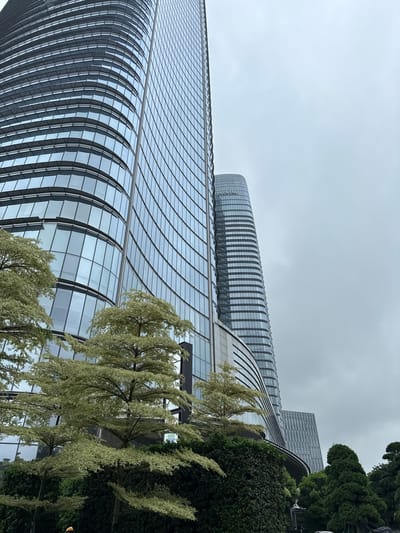 The image captures a view of modern skyscrapers in Haizhu District, China, under a cloudy sky. In the foreground, a large, curved glass skyscraper dominates the left side of the frame, its reflective facade showing a grid pattern of windows. To its right, a taller, more cylindrical skyscraper with horizontal bands is visible, bearing the text "CONTINENTAL" in large letters near its apex. The base of the structures is obscured by lush greenery, including a dense hedge and several trees with light-colored foliage and dark branches. To the far right, several conical evergreen trees are present. The overall scene suggests an urban environment with a blend of contemporary architecture and landscaping. No people are clearly visible, though a small portion of what appears to be a yellow hard hat is seen at the very bottom left corner. The lighting is diffused, indicating an overcast day.