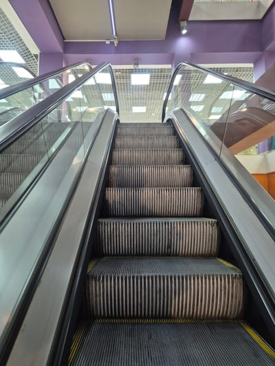 The image shows a view looking up an escalator. The escalator steps are made of dark metal with vertical ridges. The handrails are silver and made of metal with a glass panel on the side. The ceiling is a grid of white panels with lights. The walls are painted a light purple color. The scene is likely indoors, possibly in a shopping mall or public building. There are no people visible in the image. The image does not provide any specific information about the location in Petrozavodsk, Russia.