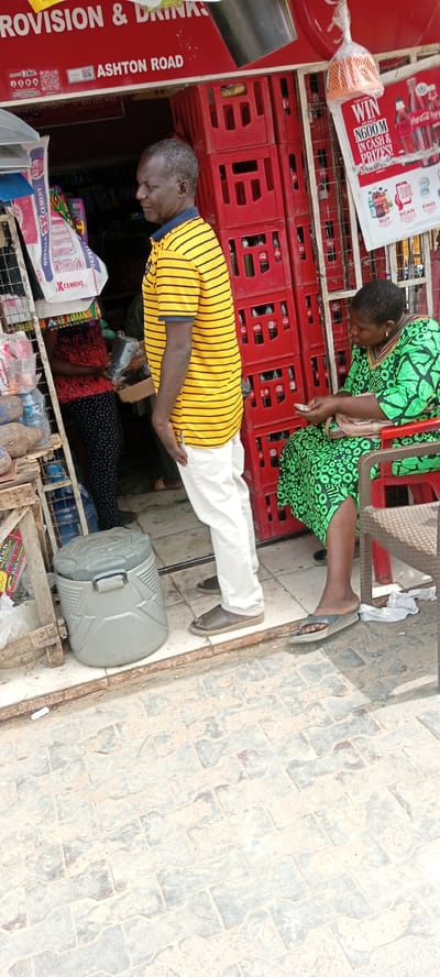 The scene is set in front of a provision store located on Ashton Road in Kwachiri, Nigeria. A middle-aged man stands near the entrance of the store wearing a yellow and black striped shirt with white pants and brown sandals. He appears to be looking towards the store's interior, where a person wearing a red and black patterned outfit is being served. Beside the man is a grey cooler. To the right, an older woman wearing a green patterned dress sits on a red plastic chair, examining paper money. Behind her are stacks of red Coca-Cola crates. A sign advertising a Coca-Cola promotional giveaway hangs on the store's facade. The ground in front of the store is paved with grey interlocking bricks. The storefront has a red sign above it, reading "PROVISION & DRINKS" and "ASHTON ROAD". Bags of cement are visible inside the store, partially obscuring the view of the person being served.