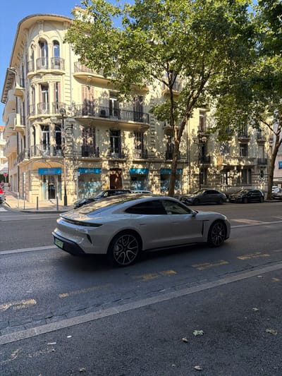 A light-colored Porsche Taycan is driving on a city street in Cannes, France. The car is in the foreground, moving from left to right. In the background, a large, ornate, light-colored building with balconies and shuttered windows is visible. Trees with green leaves are casting shadows on the building and the street. Other cars are parked on the side of the street and further down the road. The street is paved and has yellow lane markings. A few people are visible walking on the sidewalk in the distance. The sky is clear and blue.
