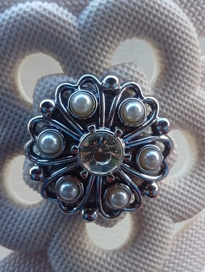 A close-up, top-down image displays a circular, silver-toned metal brooch or pin centered on a light beige, textured background. The brooch features a prominent, round, faceted clear gemstone at its core. Eight smaller, spherical, off-white pearls are symmetrically arranged around the central gem, each cradled within a curved, petal-like segment of the polished metal setting. The intricate metalwork includes small, spherical metal beads integrated into the design. The background material presents a uniform, raised waffle or cellular pattern with evenly spaced, blurred circular indentations, suggesting a soft fabric or synthetic surface. Distinct reflections are visible on the polished metal and the central gemstone, indicating a light source from the top-left relative to the object. No people, actions, or specific environmental features beyond the immediate background are discernible.