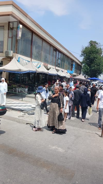 In Fergana City, Uzbekistan, a bustling street scene unfolds in front of a row of market stalls and shops. The architecture of the shops features large glass windows and a beige and brown facade, with white awnings extending over the stalls. A diverse group of people, dressed in a mix of traditional and modern attire, are walking and shopping.
Several individuals stand out: a woman in a light blue hijab and white abaya on the left, a young girl in a white dress with a blue hat, a woman in a patterned dress carrying a black purse, and a woman in a dark blue hijab and trousers. Other shoppers are visible throughout the scene, some carrying bags.
The ground is paved with asphalt and a patterned walkway. The stalls are filled with an abundance of flowers, suggesting a flower market. The weather appears to be sunny, with a clear blue sky visible above. The scene captures the everyday life and commerce of this urban environment.