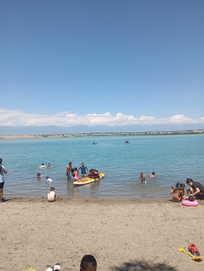 The image depicts a serene scene at Issyk-Kul Lake, a large saline lake in the Issyk-Kul Region of Kyrgyzstan, situated near the city of Karakol. The foreground shows a sandy beach with a few scattered toys, including a red toy tractor and a yellow shovel. Several people are present on the beach and in the shallow water.
In the water, closer to the shore, a young child sits facing away from the camera, looking towards the lake. Further out, a group of children are playing. One child is on a yellow surfboard, while others are in the water, some interacting with the surfboard. Two adults are also in the water near this group, seemingly supervising. On the beach, a woman in a black top and pants is attending to a child who is sitting on a pink inflatable ring. Another woman is visible in the far left of the image, facing away from the camera.
The lake itself is a vibrant turquoise, with distant mountains forming a picturesque backdrop. The sky is a clear blue with scattered white clouds, suggesting a pleasant, sunny day. The overall atmosphere is one of leisure and recreation, with people enjoying the natural beauty of Issyk-Kul Lake.