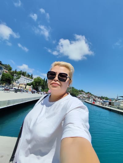 A blonde woman with sunglasses and a white t-shirt takes a selfie while standing on a pier. The woman is positioned in the foreground, with a body of blue water to her right and a sidewalk with buildings and trees behind it to her left. The sky is blue with scattered clouds. The scene appears to be a waterfront area in Sarıyer, Turkey, likely a harbor or marina given the presence of boats docked along the far bank of the water.