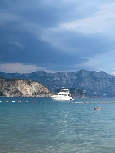 This image captures a serene scene at a coastal location, possibly in Prijevor, Montenegro. The dominant elements are the vast expanse of the sea and the imposing mountains that form a dramatic backdrop. The sea is a deep blue, with visible ripples on its surface, suggesting a calm or slightly breezy day. A white motorboat is partially visible on the left side of the frame, suggesting recreational activity.

In the water, two individuals are swimming or wading, their bodies partially submerged. One person appears to be bald and is facing the other, who is further away and facing away from the camera. They are within a marked area in the water, indicated by a line of alternating red and white buoys that extends into the distance. This buoy line likely demarcates a swimming area or a safe zone.

The mountains are rugged and rocky, with sharp inclines and shadows that suggest the sun is positioned in a way that accentuates their texture. Their scale contrasts with the human activity in the water, highlighting the grandeur of the natural landscape. The sky on the left is a deep, moody blue with darker, heavier clouds, which could indicate an approaching storm or simply a dramatic weather pattern. This contrast in the sky, with some areas appearing lighter and more diffused, adds depth and visual interest.

The overall mood of the image is one of tranquility and the enjoyment of nature. The activity of swimming in the clear waters, set against the majestic mountains, evokes a sense of peace and adventure. No text is visible in the image. The time of day appears to be late afternoon or early evening, given the lighting and cloud formations.