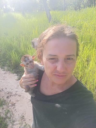 A woman is holding a baby monkey on her shoulder outdoors. The woman has fair skin, light brown hair, and wears a dark green t-shirt. Her fingernails are painted bright pink. The monkey is small and gray, with large ears and light eyes. It has a small tail and grips the woman's shoulder with its hands and feet.

In the background is a path with light-colored sand, dense tall green grass, and trees. The lighting suggests it is daytime. The location is Baan Thong Nang, Thailand.