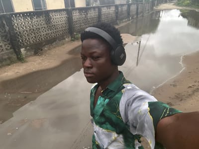 The image features a young Black person wearing black headphones and a green and white patterned shirt. They are positioned in the foreground, facing slightly to the left, with their head turned towards the camera. The scene appears to be outdoors in a wet, possibly post-rain environment, with large puddles on a sandy or dirt road. In the background, there's a textured concrete wall with a repetitive geometric pattern, and beyond that, indistinct buildings. The sky is overcast, suggesting a cloudy day. The reflection of the sky and surrounding elements is visible in the water. The overall mood is somewhat casual, possibly indicating the subject is out for a walk or enjoying music despite the damp conditions. The headphones have the "JBL" logo visible on the right earcup. The location is identified as Ajara, Nigeria.