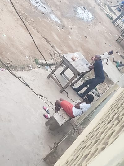 The image depicts a scene in Alalubosa, Nigeria. A man in a dark suit is leaning over a wooden table. Nearby, a woman in a white shirt and orange shorts is kneeling on a concrete slab, with a pair of sandals next to her. An electrical wire runs across the ground. Another person is visible in the background, wearing a white shirt. The surrounding environment appears to be an outdoor area with a dirt ground.