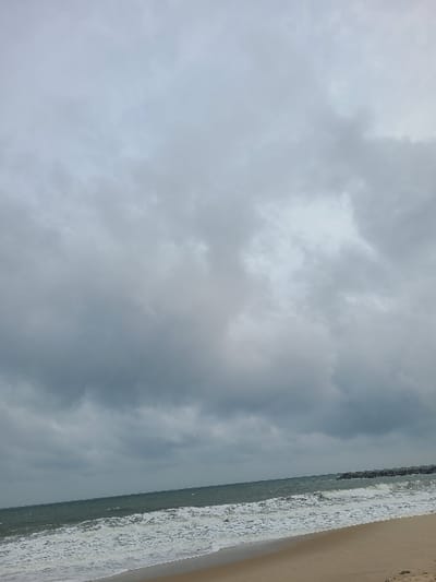 The image depicts a coastal scene under an overcast sky, presumably in Ibeju, Nigeria. Approximately two-thirds of the frame is filled with dense, grey clouds, indicating heavily overcast weather. The lower third features a body of water, likely the Atlantic Ocean, with turbulent, dark green-blue waves breaking into white foam as they approach a light brown sandy beach. A rocky breakwater or jetty, composed of large dark grey stones, extends from the right side of the frame into the water, with waves crashing against its base. No individuals or man-made structures other than the breakwater are visible. The environment appears natural and coastal, with no immediate signs of human activity on the visible beach.
