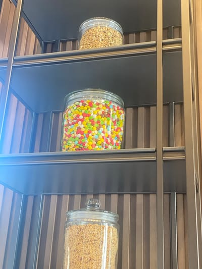 The image shows three glass jars filled with various items, arranged on shelves of a metallic structure. The top jar contains what appears to be chopped nuts or seeds. The middle jar is filled with colorful candies, predominantly M&M's or similar confectionery, in shades of red, yellow, green, blue, and white. The bottom jar, partially visible, also appears to contain chopped nuts or seeds. The shelves are made of dark material, and the vertical supports of the shelving unit are metallic and appear to be bronze or gold colored. The background behind the shelves is composed of vertical, ribbed panels in shades of brown and grey. A blueish light is cast on the left side of the image, possibly from an external light source or reflection. The scene is indoors, and no people are visible. The image is a close-up shot, focusing on the jars and the shelving unit.
The provided image does not contain any visual information that can be used to determine its location in the city of Podgorica, Montenegro. Therefore, no information about the city can be inferred from the image.