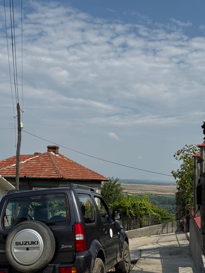 This is a vertical, full-shot photograph of a dark blue Suzuki Jimny parked on an incline. The car is facing towards the right of the frame. The rear spare tire, mounted on the back door, is clearly visible with the "SUZUKI" logo prominently displayed. The car is situated in what appears to be a residential area, with a terracotta-tiled roof visible in the lower left corner of the image. Beyond the car and the immediate surroundings, a distant landscape unfolds, featuring fields and trees under a cloudy sky. The lighting suggests it is daytime, and the presence of clouds indicates a possibly overcast or partly cloudy day. No people are visible in the image. The setting appears to be outdoors, possibly in a rural or semi-rural location. Electrical wires and a utility pole are also visible in the foreground. The overall impression is a still, everyday scene of a parked vehicle.