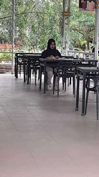 A single adult female is seated at a dark-colored square table in an outdoor dining area within Keudealue Ie Puteh, Indonesia. She wears a black hijab, a black long-sleeved top, light-colored trousers, and white patterned clogs. Her hands are positioned above a plate of food on the table, with a clear glass containing a beverage also present. The dining space features multiple dark-colored tables and chairs arranged in linear rows on a light-colored tiled floor with visible grout lines. The area is covered by a structure supported by white metal poles and beams. The background consists of dense green foliage. A small sign depicting various drinks is attached to a pole in the upper right quadrant of the image. A white motorcycle is partially visible in the distant background on the right.