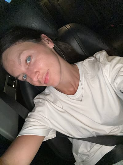 A woman with light blue eyes and fair skin is seen from a close-up perspective, looking upwards towards the camera. She is wearing a white t-shirt and a thin silver necklace with a small pendant. Her hair is dark brown and appears to be pulled back. She is seated in a car, indicated by the presence of a black leather seat and a seatbelt across her chest. The lighting suggests it is either nighttime or a dimly lit interior. There are no discernible urban features or landmarks to indicate a specific location within the city of Boreti, Montenegro.