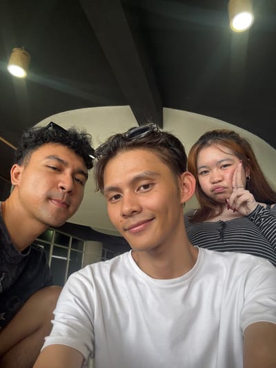 Three people are posing for a selfie indoors, likely in a cafe or restaurant in North Jakarta, Indonesia, given the presence of a pendant lamp and a black ceiling with visible beams, common in such establishments.
In the foreground, a man with curly dark hair and sunglasses perched on his head is on the left, looking directly at the camera with a slight smile. He is wearing a dark t-shirt. Next to him, a man with light brown hair and sunglasses on his head is in the center, also smiling at the camera. He is wearing a white t-shirt. To the right, a woman with long, light brown hair is making a pouty face and holding her hand up to her cheek. She is wearing a black and white striped shirt. The background shows a modern interior with large windows and a dark ceiling with light fixtures.