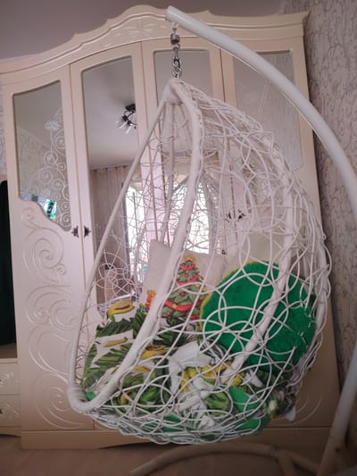 This is a close-up, medium shot of a white wicker hanging chair, with pillows inside, in a room. The chair is suspended from a white metal frame. The pillows have a tropical leaf pattern, a Christmas tree pattern, and a plain green pillow. In the background, there is a cream-colored wardrobe with mirrored doors that reflect a ceiling light fixture. The wardrobe has decorative carvings. The floor appears to be wooden, and the wall has a patterned wallpaper. There is no indication of the city of Hrodna, Belarus, in the image or its metadata.
