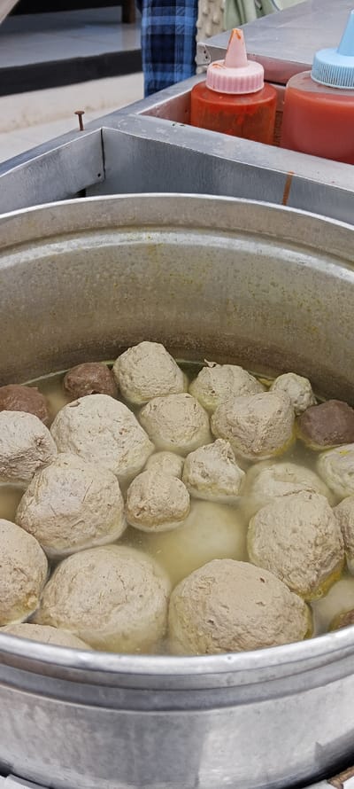 The image shows a large pot of meatball soup, located at coordinates -6.973, 107.52 in Kecamatan Kutawaringin, Indonesia. The pot is made of aluminum and contains approximately 15 meatballs. The soup is a light brown color, and the meatballs are all round and smooth. Two plastic bottles of sauces are visible in the background, one red and one blue. One bottle contains a red sauce and has a pink lid, the other bottle contains red sauce and has a blue lid. There is a rusty nail on the side of the pot.  The background of the photo shows the leg of a person wearing a plaid shirt.  The scene appears to be outside, possibly a street food stall.