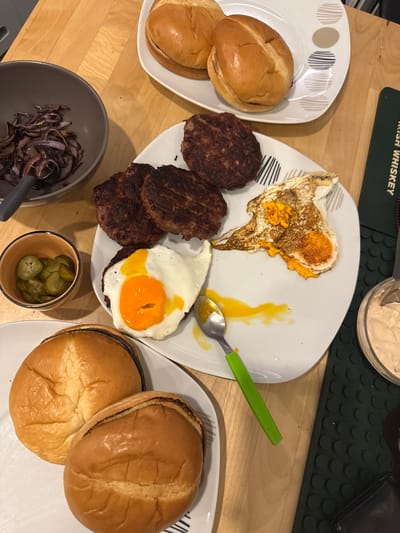 The image shows a top-down view of a meal laid out on a wooden table. The meal consists of hamburger buns, beef patties, a fried egg, sliced pickles, and what appears to be caramelized onions. The food is arranged on white plates, with a small bowl of pickles and a larger bowl of onions to the left. A green-handled spoon is resting on one of the plates next to the fried egg. To the right, a jar of sauce and a green coaster are visible. The overall scene suggests a home-cooked meal. The image does not contain any information that can be used to determine its location in Almada, Portugal.