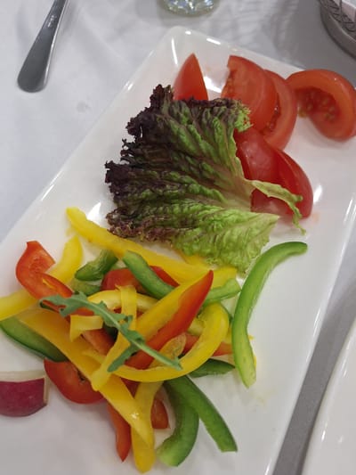 A white rectangular plate sits on a white tablecloth, presenting a colorful assortment of sliced vegetables and a leaf of dark red lettuce. The plate contains several wedges of ripe red tomatoes, their seeds and juicy interiors visible. Interspersed with the tomatoes are sliced bell peppers in shades of yellow, green, and red, cut into thin strips. A single leaf of dark red lettuce, with ruffled edges and green veins, rests prominently on top of the bell peppers. Some arugula leaves are scattered amongst the peppers, adding a touch of green. A sliver of a red-skinned vegetable, possibly a radish, is visible at the bottom left edge of the plate. A silver fork is partially visible on the upper left side of the image, positioned on the tablecloth. The scene is lit by overhead light, creating subtle shadows and highlighting the textures of the food. The background is out of focus, suggesting an indoor dining setting. The context of this image being in Peterhof, Russia, implies it may have been taken at a restaurant or a private residence within the city, possibly near one of its famous palaces or gardens.