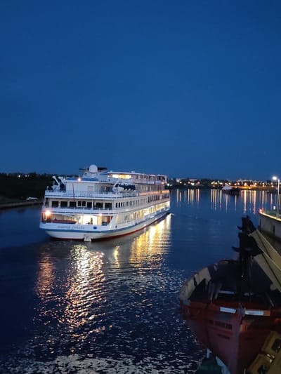 A large white cruise ship, named "ANDREY RUBLEV" in Russian, travels down a calm body of water at dusk. The ship has multiple decks with illuminated windows and is casting reflections on the water. In the foreground, a smaller red boat with dark railings is partially visible, also in the water. The sky is a deep blue, transitioning to darker hues as night approaches. In the background, the lights of a distant city or town are visible along the shoreline, along with trees. A street lamp is also visible on the right side of the frame. The scene appears to be a waterway in or near the city of Sheksna, Russia, possibly a canal or river given the urban and natural elements.