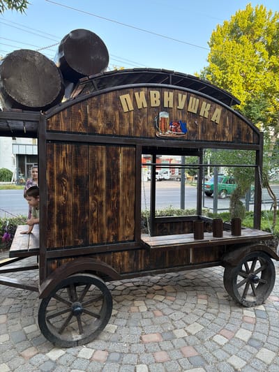 A wooden cart with a rustic design, resembling a beer stall or kiosk, is parked on a cobblestone pavement in Chirchik, Uzbekistan. The cart is adorned with large, dark barrels on its roof and a sign that reads "ПИВНУШКА" (Pivnushka), which translates to "Beerhouse" or "Little Beerhouse" in Russian. Below the sign, there is a graphic of a beer mug and some food items.
A young boy, wearing a light green shirt, is leaning over the counter of the cart, appearing to be engaged with something outside the frame. A woman, dressed in a patterned shirt, stands behind him, partially visible. The cart features wooden planks for its walls and shelves, and large, spoked wheels that give it a vintage appearance.
In the background, across a street, there are buildings, trees, and other vehicles, including a distinctive green Soviet-era car. The sky is clear and blue, indicating fair weather. The overall scene suggests an outdoor setting, possibly a park, a street market, or a public area in the city.
