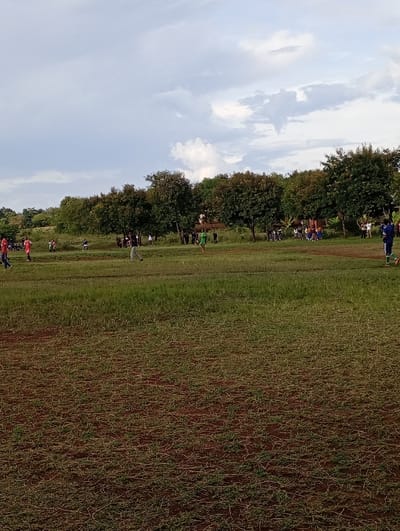 The image shows a grassy field with a few people on it. It appears to be a soccer field located in Arusha, Tanzania, near the coordinates -3.438 latitude and 36.733 longitude.  The field has short, green grass and a few patches of bare, brown soil. The field is surrounded by trees and bushes. There are a few people in the distance, some wearing soccer uniforms and some in casual clothing. Some are walking on the field while others seem to be watching the game. The sky is overcast, but there are a few patches of blue sky visible.