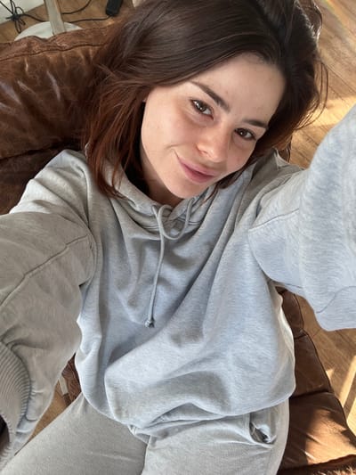 The image depicts a close-up, high-angle selfie of a single female subject in an indoor residential setting. The subject, a young adult with light brown hair, visible freckles across her nose and cheeks, and brown eyes, is positioned reclining on a distressed brown leather object, likely a sofa or armchair. She is attired in a light grey hooded sweatshirt with visible drawstrings and matching light grey sweatpants. Her facial expression exhibits a slight smile, with her gaze directed towards the camera. Strong natural light, originating from the right side of the frame, illuminates a wooden floor visible in the background and casts distinct shadows. No specific environmental markers or architectural features are discernible within the image to identify a precise location within the city of Roubaix, France.