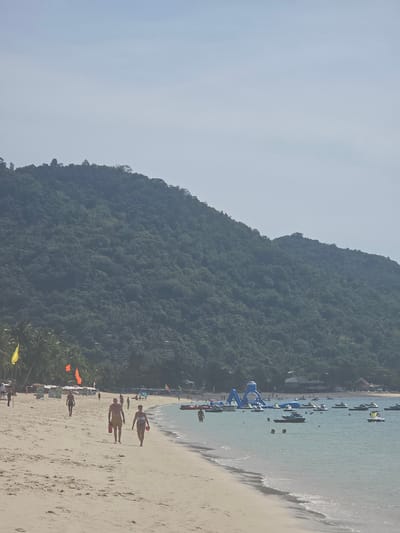 The image depicts a tropical beach scene in Baan Lamai, Thailand. A wide, sandy beach occupies the foreground, with people walking along the shoreline. In the distance, the clear blue water is dotted with numerous jet skis and a large inflatable water park structure. Lush, green, tree-covered mountains form a dramatic backdrop to the beach. The sky is a pale blue with a slight haze. Several colorful flags and umbrellas are visible on the beach, suggesting a public tourist area. A few palm trees are scattered along the edge of the sand.