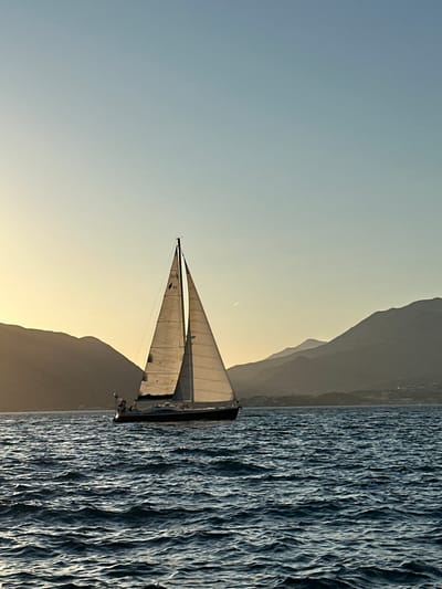 A sailboat navigates the waters off Luštica, Montenegro, captured during either sunrise or sunset. The vessel, centrally positioned but slightly right, features a dark-colored hull and two prominent triangular sails, which are illuminated by a warm, low-angle light originating from the left. Faint numerical markings, "02" and "50," are discernible on the forward sail. The surrounding sea exhibits dark blue, choppy waters with distinct ripples, indicating movement. In the background, multiple layers of dark, silhouetted mountains span the horizon, receding into atmospheric haze. The sky transitions from a soft golden-yellow glow on the left, near the light source, to a pale blue hue in the upper and right portions of the frame. A single, thin white contrail is visible high in the sky to the right of the sailboat. Distant, indistinct structures are present along the coastline at the base of the mountains on the far right. No discernible human figures are clearly visible aboard the boat.