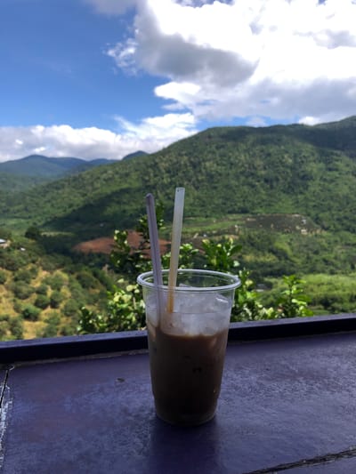 A close-up, eye-level shot focuses on a plastic cup of iced coffee sitting on a dark purple, weathered surface. Two straws, one purple and one translucent white, emerge from the coffee, which is a light brown color with ice cubes. In the background, lush green mountains rise under a bright blue sky dotted with white clouds. The mountains are covered in dense vegetation, with some areas appearing to be terraced or cleared. The overall scene suggests a scenic overlook in La Dạ, Vietnam, likely a cafe or viewpoint.