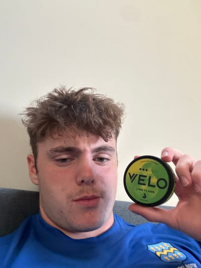 A man with curly brown hair and blue eyes holds a green and yellow can of Velo Lime Flame nicotine pouches. He is wearing a blue t-shirt with a patch. The background is a plain, light-colored wall. He is looking directly at the camera with a neutral expression. The image is slightly rotated.