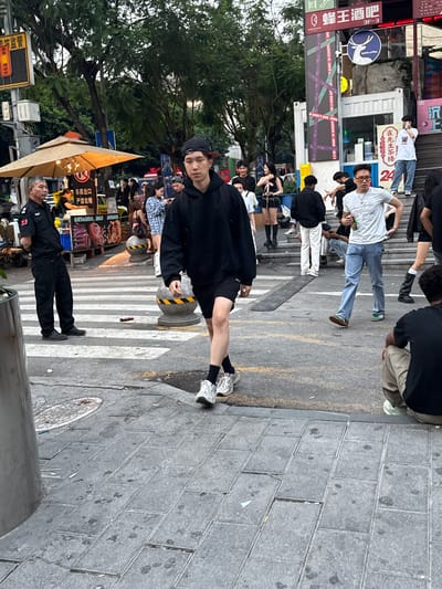 The image depicts an outdoor scene in Liangjiang, China, populated by multiple individuals in a commercial area with various shops and signage. In the foreground, a young person wearing a black hooded sweatshirt, black shorts, white socks, and sneakers stands on a crosswalk. An older individual in a black security uniform stands nearby. The environment includes street signs, an umbrella-covered food stall, a "luckin coffee" sign, and other commercial signage in Chinese. In the background, pedestrians mill about, and there's a crosswalk with alternating white and black bollards. The ground consists of gray, brick-like pavers.