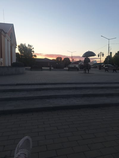 The image depicts a dusk scene in the city of Kapyĺ, Belarus. The sky is a soft gradient of blue and pink, indicative of sunset. In the foreground, a person's foot in a grey and white athletic shoe is visible, suggesting the image was taken from a low vantage point.
The middle ground features a paved square with rows of dark grey brickwork, leading up to a series of raised steps. Beyond the steps, a public space is visible. To the left, a building with arched windows and columns, possibly a public institution, is partially seen. In the center of the square, a bronze sculpture of two figures holding an umbrella stands prominently. Two dark benches are placed on either side of the sculpture, and further in the background, streetlights and trees are visible, along with other buildings. The overall atmosphere is quiet and serene, consistent with early evening in a city square.