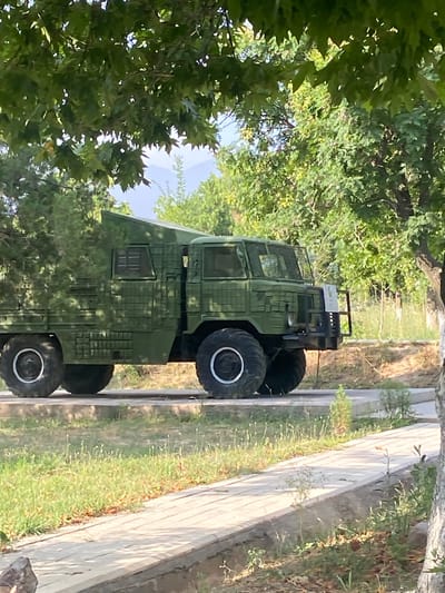 This is a photograph of a large, olive-green, military-style vehicle parked outdoors. The vehicle has prominent, rugged tires and a boxy, utilitarian design with multiple windows. It appears to be an off-road truck or transport vehicle. The background consists of lush green trees and foliage, suggesting a natural or park-like setting. A light-colored pathway or sidewalk runs along the right side of the image, bordered by grass and dry vegetation. The lighting indicates it is daytime, likely sunny, given the shadows and highlights. The scene is static, with no visible activity or people. The image is oriented vertically, with the vehicle appearing on its side in the composition. There is no visible text in the image. The context places this scene in Angren, Uzbekistan.