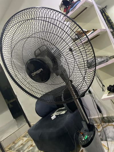 A close-up shot focuses on a black electric fan with a dense, wire mesh guard. The fan's central hub displays the brand name "GASA." A metallic pole extends from the fan base towards the top right, where it connects to the fan's motor. The base rests on what appears to be a tiled floor. In the background, partially visible are items on shelves, a black bag, and power cords, suggesting a cluttered indoor environment.