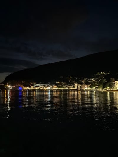 The image depicts Petrovac centar, Montenegro, at night, from the perspective of the sea. The dark sky is partially obscured by faint cloud formations, visible above a prominent, unlit mountain that forms a dark silhouette spanning the upper half of the background. Along the base of the mountain, a densely built coastal town is illuminated by numerous artificial lights. These lights, originating from buildings, streetlights, and possibly commercial establishments, cast bright reflections across the dark, rippled surface of the water in the foreground. A distinct, intense blue light emanates from a structure on the left side of the town, creating a prominent blue reflection on the water. Other lights appear yellow or white, contributing to a tapestry of broken light streaks on the water's surface. No individuals or specific activities are discernible. The overall scene conveys a nocturnal urban coastal landscape with significant light pollution reflecting on the water.