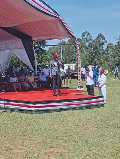 The image shows a man in a black suit and a red hat standing on a red stage, speaking into a microphone. The stage is under a white and red tent with black trim. The stage is surrounded by about 20 people sitting on chairs on a green grass field. The people are mostly men. The man in the black suit appears to be giving a speech. The scene is outdoors and the sky is blue with white clouds. There is a green leafy background. The scene is in Malava, Kenya, located at latitude 0.433, longitude 34.907.  There is also a large white truck partially visible in the background, to the right of the tent.  Several people are standing near the truck. Two people are standing to the right of the stage, one in a white shirt and white pants, and one in a white shirt, blue pants, and a blue cap.  The man speaking appears to be the main focus of the image.  There is also a microphone stand in front of the stage.