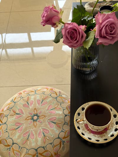 A top-down shot shows a surface with a vase of pink roses, a teacup filled with dark liquid, and a round patterned cushion, with part of a light-colored tiled floor visible below. The setting is Givatayim, Israel.