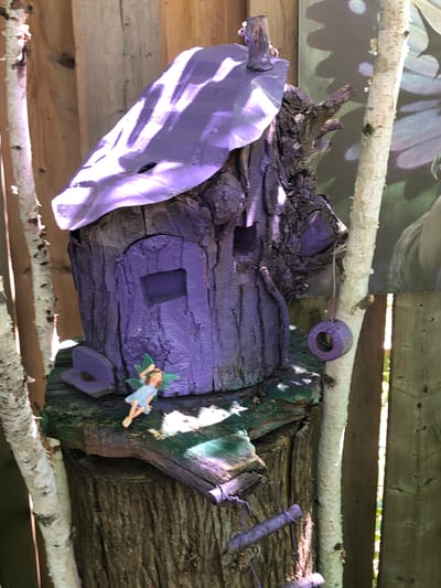 The image captures a whimsical, handcrafted fairy house situated in a garden setting. The house is constructed from a hollowed-out log, painted a vibrant purple. It features a purple roof with a rough, uneven texture, and a small, rectangular window. A miniature fairy figurine, dressed in a light blue dress, stands near the base of the house, appearing to be interacting with the structure.

The fairy house is perched atop a weathered tree stump, which serves as its foundation. Surrounding the stump are remnants of painted wood, some green and others a darker hue, suggesting it's part of a larger garden decoration or play area. To the right of the house, a small, purple sphere hangs from a string, possibly a decorative element or a miniature bird feeder.

In the background, a wooden fence with vertical planks provides a rustic backdrop. A large, abstract artwork or mural, featuring floral or leaf-like patterns in shades of green and purple, is partially visible against the fence. Two slender, white-barked birch trees frame the fairy house on either side, their branches reaching upwards. Sunlight filters through the leaves, casting dappled shadows across the scene, indicating an outdoor, possibly daytime, setting.

This particular fairy house is located in the city of Severn, Canada.