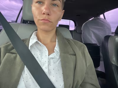 A person is in the driver's seat of a car, wearing a white shirt with black polka dots, a light green jacket, and a seatbelt. A white bag is behind the person's head, suspended from the headrest. The exterior view out the window suggests the location is in Verkhniy Mamon, Russia, with the sky colored purple and power lines visible.