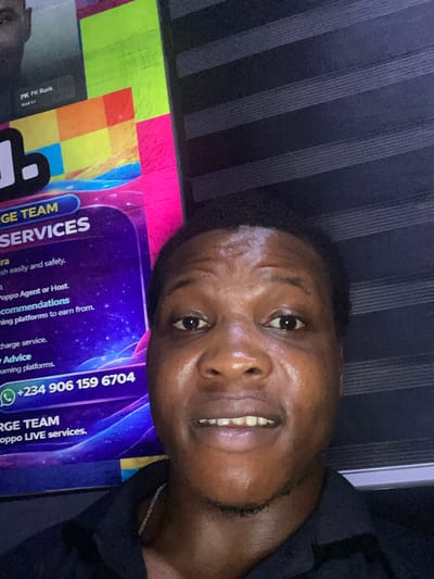 The image shows a person, likely a young adult male, facing the camera with a slight smile. He has short, dark hair and appears to be of African descent. His skin is slightly shiny, possibly due to perspiration. He is positioned in front of a poster or sign. The poster features the words "GE TEAM SERVICES" and includes text related to the Poppo app, suggesting it's a marketing or promotional material for live streaming or earning opportunities. The poster also displays the phone number +234 906 159 6704. A wall is visible behind the person and poster. Based on the phone number with the country code +234, it can be inferred the location is Nigeria, and given that there are services advertised on the poster, it is likely the scene is in a commercial location. No definitive clues indicate the precise city or specific location such as Alor.