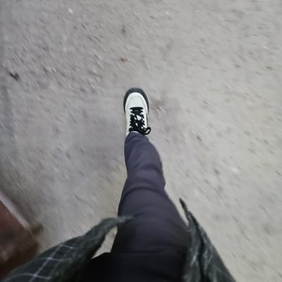 The image shows a downward-angled view of a person walking on a grey concrete surface, likely a sidewalk. The individual is wearing dark trousers and a plaid jacket. The focal point is a white sneaker with black laces and a black sole, suggesting movement. The background is blurred, indicating motion. The location is not apparent, and without visual landmarks or other context, it's impossible to ascertain the specific location within Fez, Morocco.