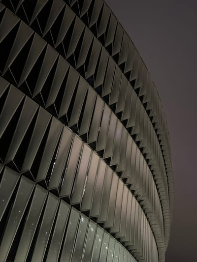 The image shows a close-up, upward-angle view of the exterior of the San Mamés Stadium in Bilbao, Spain. The stadium's facade is characterized by a repeating pattern of angled, dark gray metallic panels that create a textured, faceted surface. These panels are arranged in rows that follow the curve of the stadium's structure. Some of the panels appear to be translucent or have lights behind them, casting a diffused glow and reflecting small points of light. The sky above is a muted, dusky gray, suggesting it is nighttime or twilight. The overall impression is one of modern, architectural design with a focus on geometric patterns and material texture, highlighting the stadium's distinctive external appearance. There are no people or other discernible objects in the image.