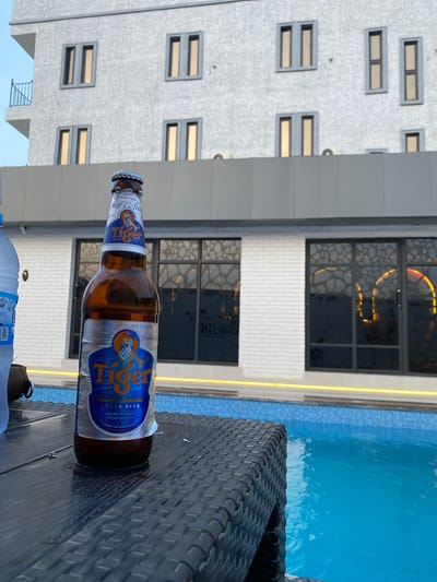 The image, taken in Kubwa, Nigeria, captures a tranquil poolside scene during what appears to be late afternoon or early evening. In the immediate foreground, a bottle of Tiger beer rests on a dark, woven outdoor table or chair. The beer bottle's label is prominent, featuring the brand name "Tiger," along with text such as "LAGER BEER," "AWARD WINNING," "PREMIUM BEER," and "SINGAPORE SINCE 1932," indicating its origin and quality. A portion of a plastic water bottle is also visible on the table.
Beyond the table, a clear blue swimming pool with tiled edges reflects the bright sky and surrounding structures, suggesting calm water. The background is dominated by a modern, multi-story building. Its lower section features a white, brick-textured facade with large dark windows that have intricate, possibly illuminated, patterns inside. The upper floors of the building are light grey, punctuated by numerous small, rectangular windows that emit a warm, golden glow from within. The overall setting suggests a leisurely environment, possibly a hotel, resort, or an upscale residential complex. The sky is bright and clear, contributing to the relaxed ambiance of the scene.