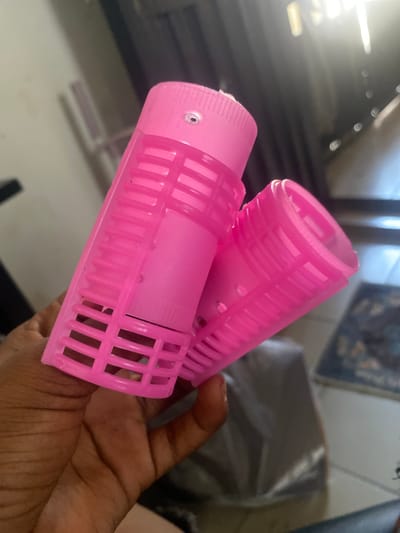 In Kubwa, Nigeria, a hand holds two bright pink plastic hair rollers. The rollers are cylindrical with a slotted design. One roller is positioned vertically, while the other is angled in front of it, partially obscuring it. The background is blurred, showing hints of indoor lighting and possibly furniture. The image is taken from a close-up perspective, focusing on the hair rollers and the hand.