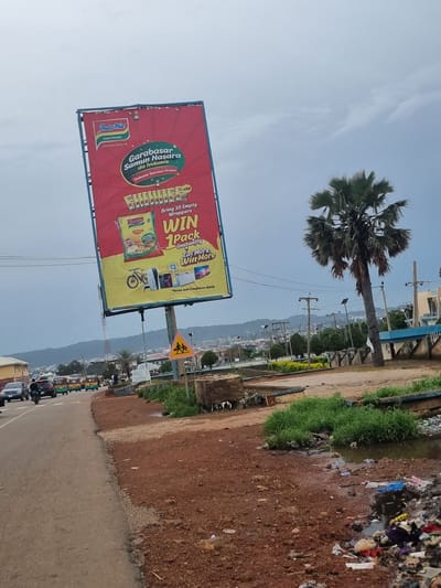 The image shows a street scene in Rafin Par, Nigeria, dominated by a large billboard advertising Indomie instant noodles. The billboard, tilted at an angle, features a promotional message about winning prizes by bringing empty noodle wrappers. Various prizes like a bicycle, a fan, and a television are depicted.
The street itself is paved, with a dirt shoulder on the right side. Several vehicles, including cars, a motorcycle, and tricycles (popularly known as "Keke Napep"), are visible on the road, suggesting traffic. The environment appears somewhat neglected, with piles of garbage and debris accumulating near the roadside, particularly on the right. Lush green grass grows in patches, contrasting with the dry earth and accumulated waste. In the background, a palm tree stands prominently, and a distant cityscape with buildings and hills can be seen under an overcast sky. Power lines and utility poles are also present, indicating the presence of infrastructure. A yellow warning sign with a symbol of two people crossing is visible near the billboard. The overall impression is a typical urban or semi-urban environment in Nigeria, marked by commercial advertising, transportation, and the challenges of waste management.