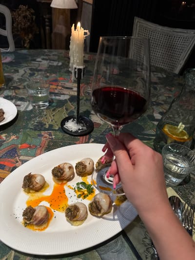 A high-angle, indoor shot depicts a dining experience. An oval, white plate displays escargots, garnished with herbs and bathed in an orange-yellow sauce. A hand with pink nail polish reaches for a snail fork resting within one of the shells. A glass of red wine lies horizontally on the table alongside a candle in a black candlestick, a water pitcher with citrus, and a glass of water. Silverware is placed at the top of the scene. The tablecloth has a patterned design. The location and city cannot be determined from the given image.