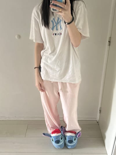 This full-length, vertical image depicts an individual standing indoors, reportedly in Cachan, France, facing a reflective surface while holding a smartphone. The individual, possessing long dark hair, is dressed in a white short-sleeved t-shirt displaying a light blue New York Yankees logo and a smaller MLB logo, light pink sweatpants with a visible drawstring, and a pair of blue "Stitch" character slippers, each wearing a small red and white Santa hat. A ring is visible on the right index finger, and dark beaded bracelets are present on both wrists, with an additional black spiral bracelet on the right wrist. The background features a plain, light-colored wall with a circular white fixture on the left side. The floor is light-colored, possibly wood or laminate, and a vertical white door frame or mirror edge is visible on the right.