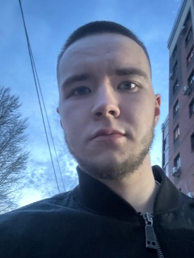 A selfie image shows a person with short, dark hair and a beard, wearing a dark-colored hooded jacket. The background features a building with multiple windows and a sky with clouds and bare tree branches. Electrical wires are visible against the sky. The location cannot be verified.